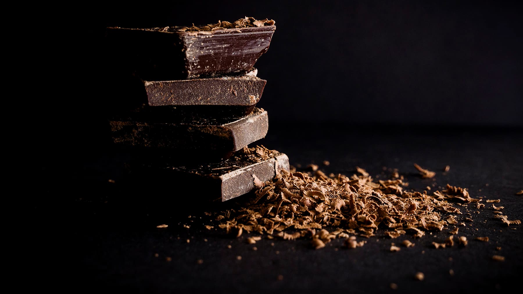 Stacked dark chocolate bars with chocolate shavings on a dark surface.